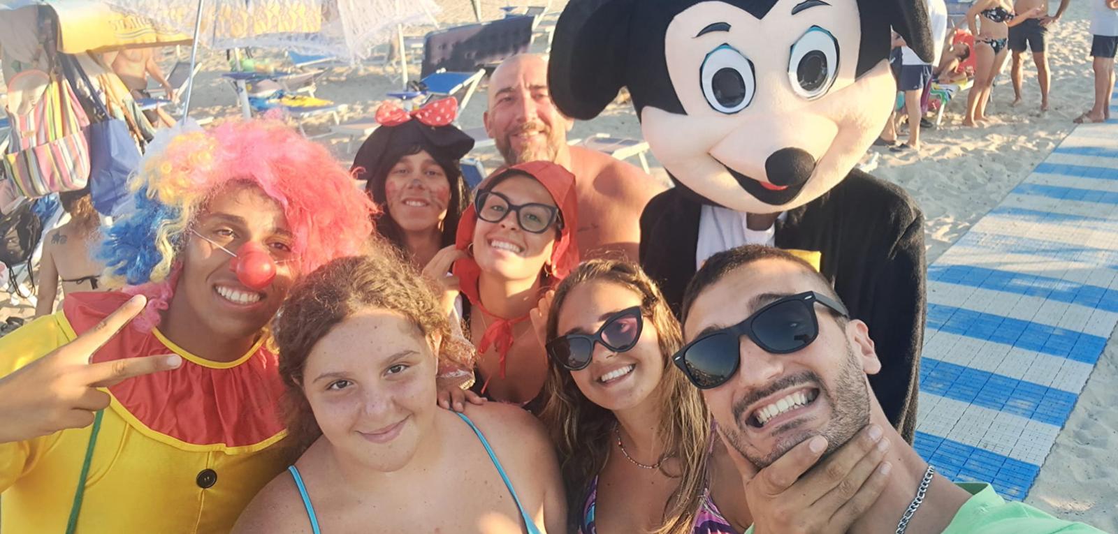 Gruppo di persone in spiaggia con costumi colorati e un personaggio famoso.