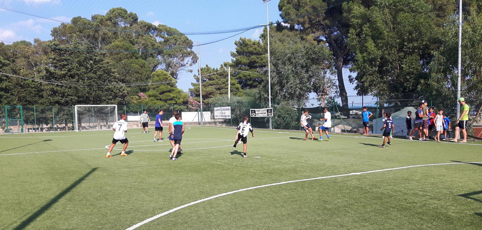 Partita di calcetto tra ragazzi in un campo all'aperto, con spettatori a bordo campo.