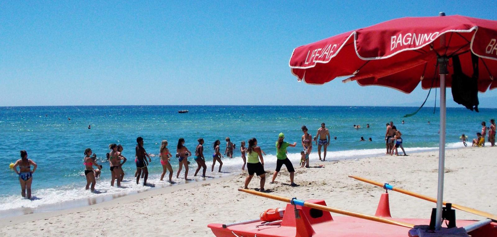 Spiaggia soleggiata con persone che fanno esercizi vicino al mare, ombrellone e pattino di salvataggio.