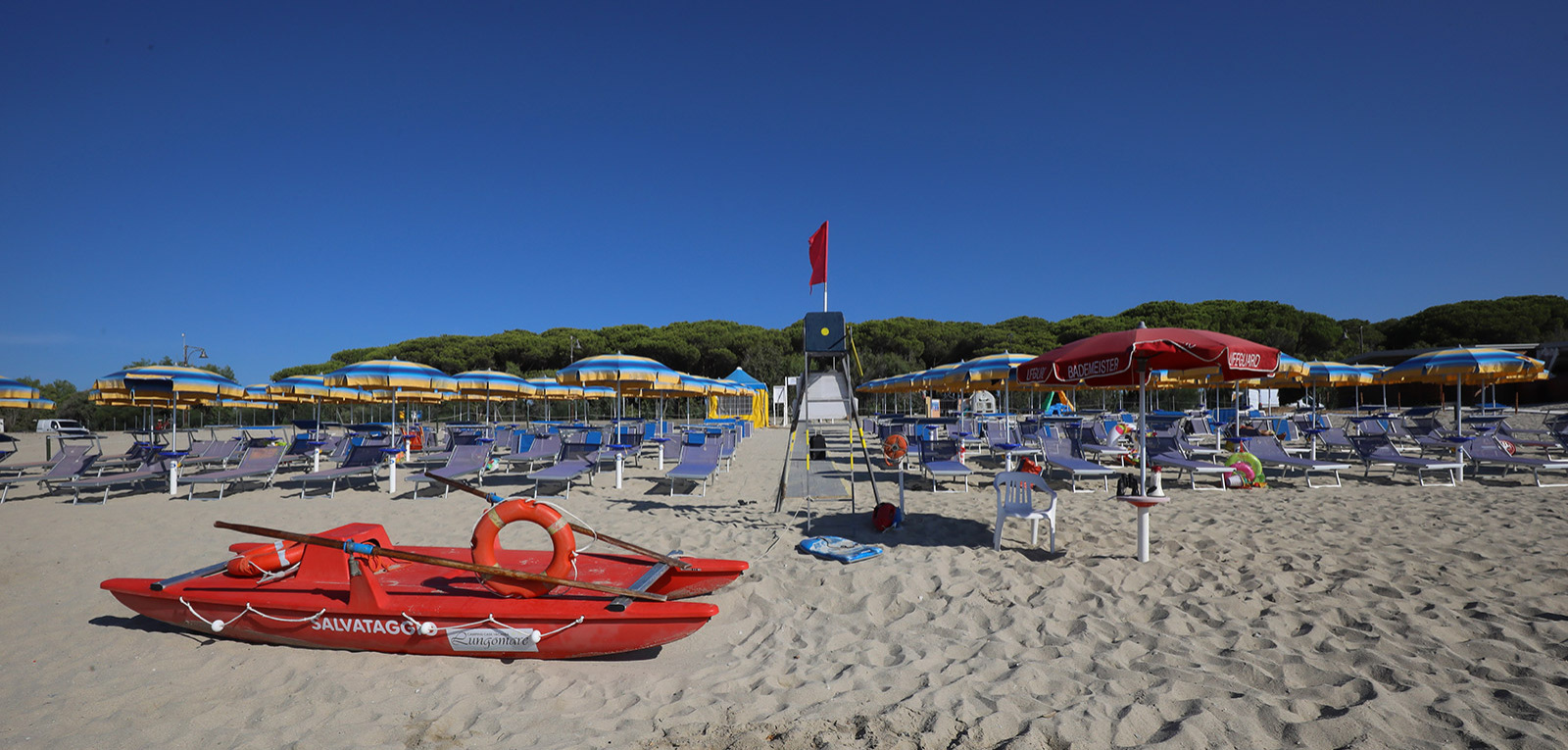 Spiaggia con ombrelloni gialli e blu, lettini e un pattino rosso per salvataggio.