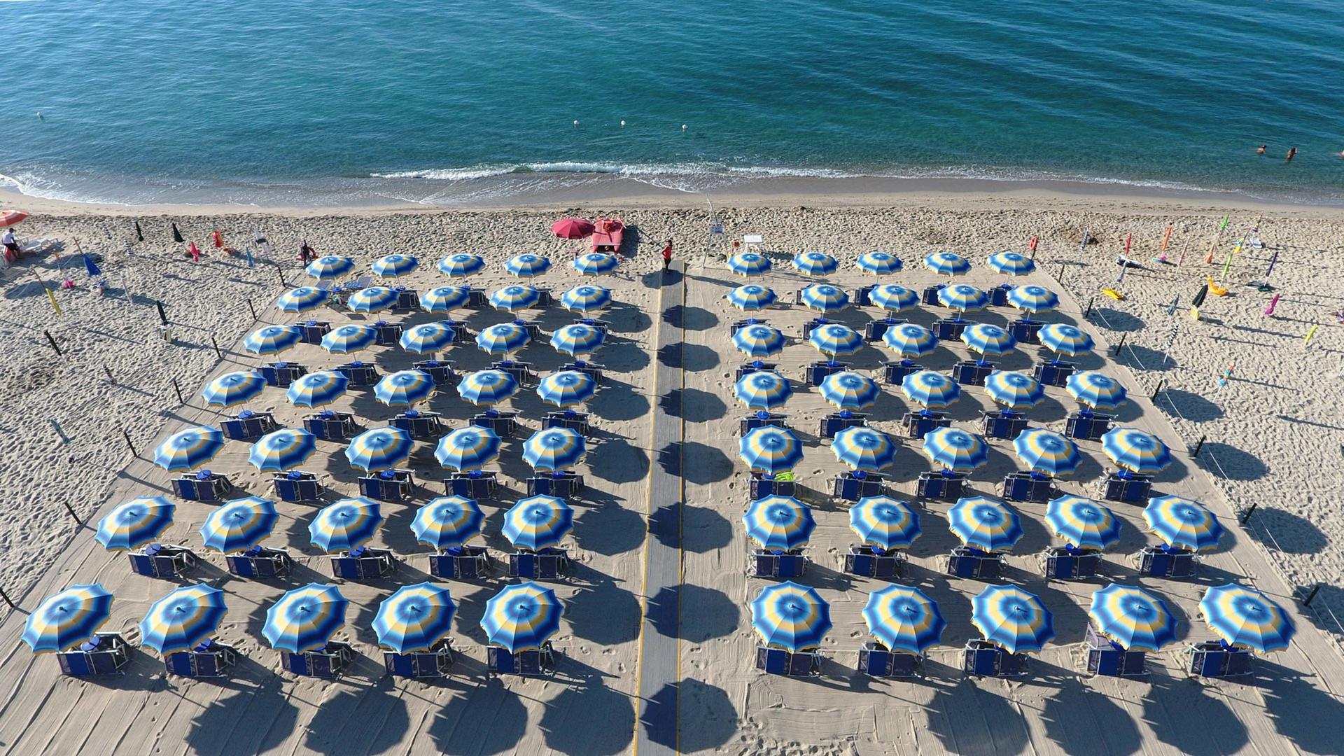 Spiaggia affollata con bagnanti e ombrelloni, mare cristallino e cielo sereno.