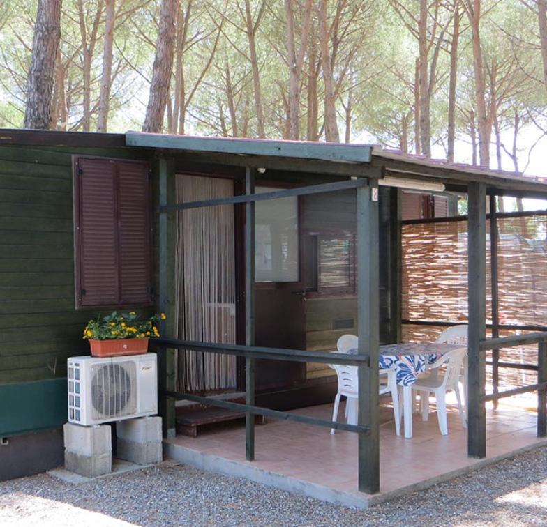 Bungalow in legno con veranda coperta, tavolo e sedie, immerso in una pineta.